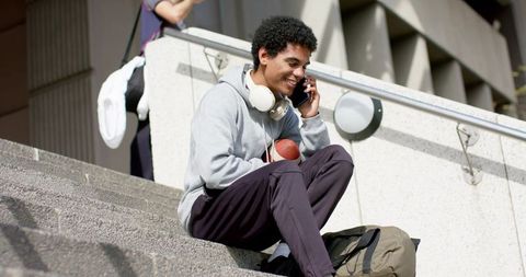 African american college student sitting on steps talking on phone while holding football