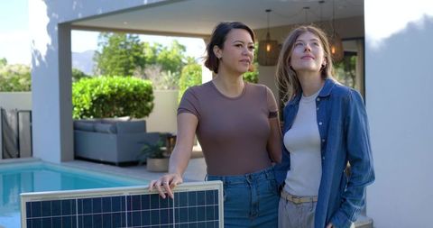 Two women admiring solar panel on modern backyard patio with pool, greenery and sustainable living