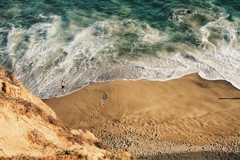 Aerial coastal cliff overlooking turquoise surf with lone runner leaving footprints on sand