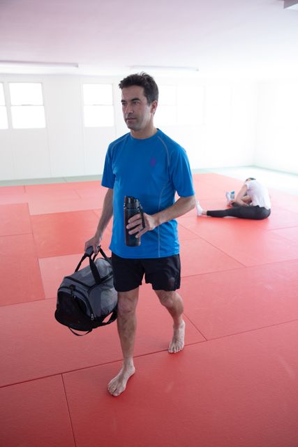 Caucasian male judo coach holding a gym bag and plastic flask, leaving the gym after a training session. A female judoka is stretching in the background on a red mat. This image can be used for promoting martial arts training, fitness programs, sports equipment, or gym facilities.
