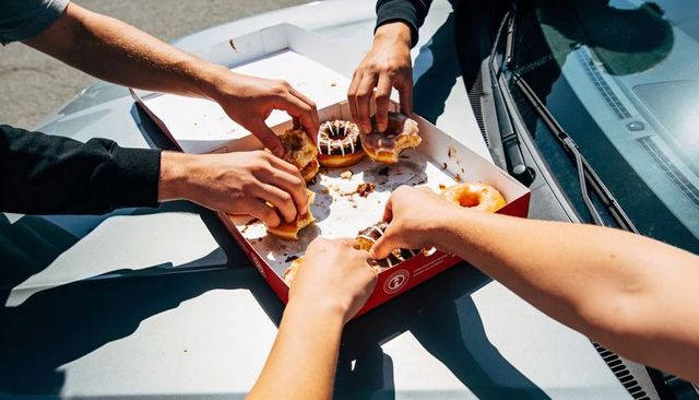 Friends sharing donuts on car hood during sunny road trip reaching into red box
