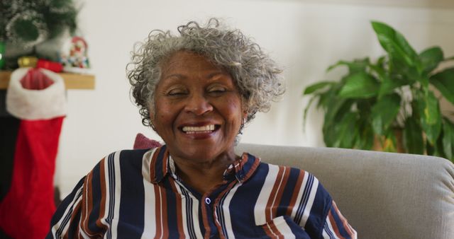 Senior African American woman wearing striped shirt, cheerful and smiling, sitting in living room with Christmas decorations and indoor plant in background. Suitable for celebrating the holiday season, senior lifestyle, happiness, and family gatherings.