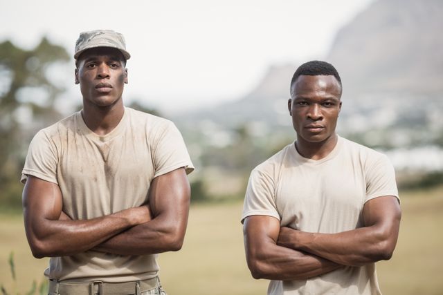 Two African American military soldiers standing with arms crossed during an outdoor boot camp. They are wearing uniforms and appear serious and determined. This image can be used to depict themes of military training, discipline, teamwork, and physical fitness. Ideal for articles, advertisements, or websites related to military life, fitness programs, or team-building activities.