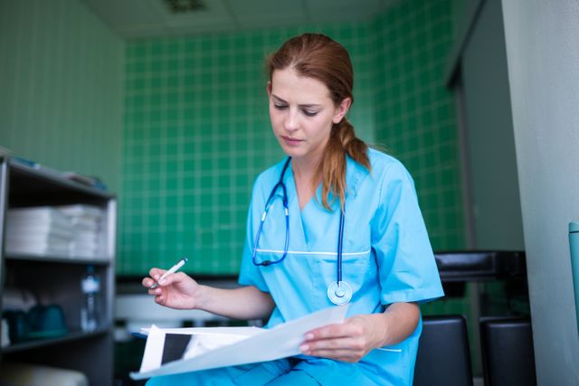 Nurse in blue uniform with stethoscope around neck reviewing medical report in hospital. Ideal for use in healthcare, medical, and hospital-related content. Can be used in articles, brochures, and websites focusing on nursing, patient care, and medical professions.
