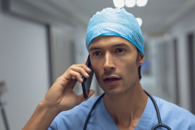 Male surgeon in blue scrubs and cap talking on mobile phone in hospital corridor. Stethoscope around neck. Useful for healthcare, medical communication, emergency response, hospital staff, and healthcare industry themes.