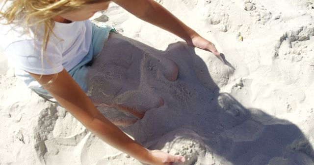 A young Caucasian woman is enjoying a playful moment at the beach, sculpting sand with her hands, with copy space. Her creativity is showcased by the sand figure she's crafting, capturing the essence of summer fun.