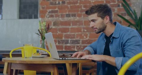 Young man typing on laptop at cafe with orange juice and pastry, denim shirt, exposed brick