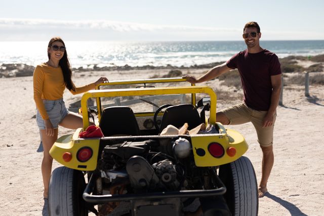 Portrait of happy caucasian couple standing by beach buggy on sunny beach by the sea. beach stop off on romantic summer holiday road trip.