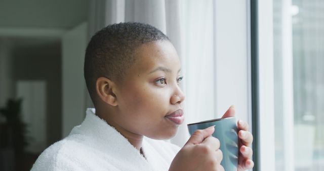 Thoughtful african american plus size woman wearing robe, looking through window, drinking coffee. domestic lifestyle, enjoying leisure time at home.