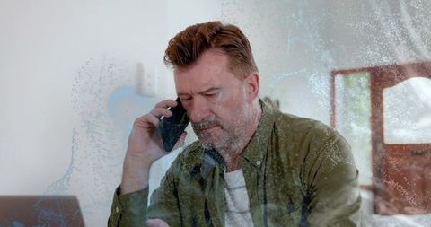 Midlife man working remotely from home on phone and laptop through window glass with guitar
