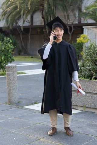 Young asian graduate wearing cap and gown talking on phone holding diploma outdoors