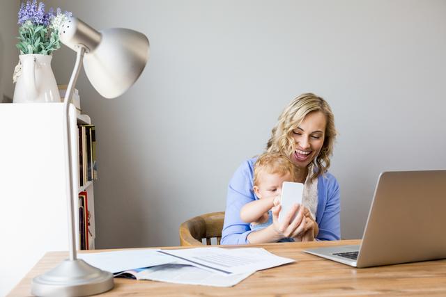 Mother and baby sitting at a desk in a home office, with the mother holding a mobile phone and smiling. The baby is on her lap, engaging with the phone. A laptop, documents, and a lamp are on the desk. Ideal for use in articles about work-life balance, parenting, remote work, and modern family life.