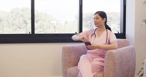 Nurse in pink scrubs checking watch while using phone in clinic waiting area