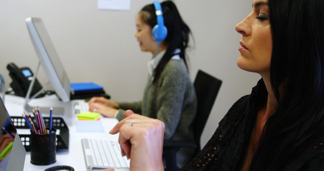 Two women working at their desks in a modern office setting. The woman in the foreground is focusing intently on her task, while the woman in the background is wearing headphones while using a computer. Ideal for illustrating concepts related to workplace productivity, teamwork, modern office environments, or gender diversity in the workplace.