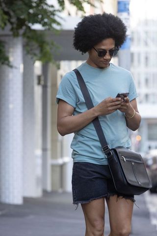African american man walking in city checking smartphone with messenger bag and sunglasses