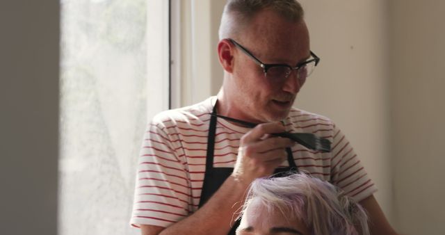 Hairdresser is coloring hair with brush, wearing glasses and apron. Modern hair salon setting with natural light through window, creating professional and welcoming environment. Suitable for illustrating beauty services, hairstyling process, hair care marketing materials, or salon promotions.