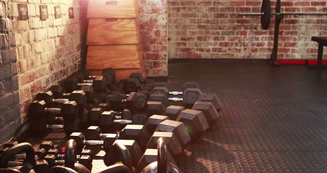 A variety of dumbbells are scattered on the floor of a gym with brick walls and exercise equipment in the background. The setting suggests a robust workout environment where individuals focus on strength training and physical fitness.