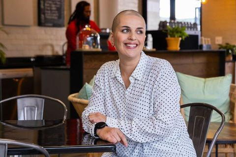 Smiling bald woman sitting at glossy cafe table wearing smartwatch, cozy modern coffeehouse