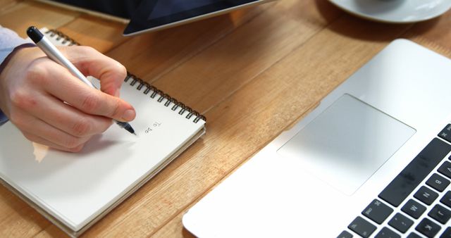 Close-up of person writing notes in spiral notebook on wooden desk with open laptop nearby. Useful for topics related to productivity, studying, business planning, or remote work. Perfect for illustrating home office setups, study sessions, or organizational strategies.