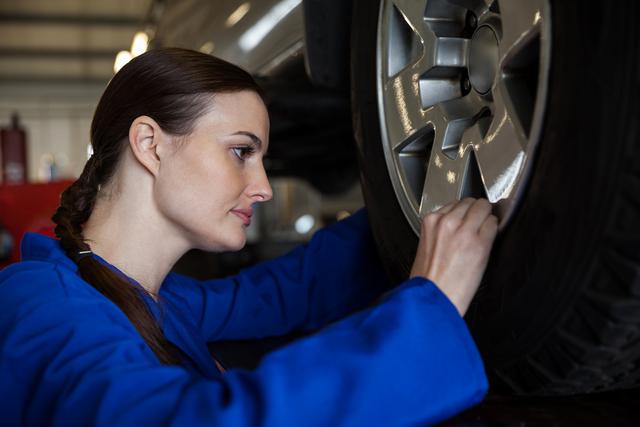 Image shows a female mechanic inspecting a car wheel in a repair garage. Ideal for illustrating automotive services, professional mechanics at work, car maintenance, and technical skills in women. Useful for articles, advertisements, and marketing materials related to car repair shops, automotive engineering, and gender diversity in technical fields.