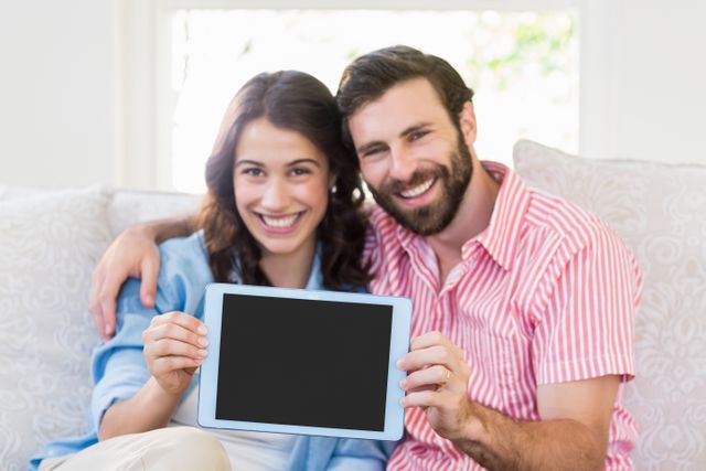 Couple sitting on sofa in living room, smiling and holding digital tablet with blank screen. Ideal for technology, family lifestyle, and home relaxation themes. Can be used in advertisements for tech products, family-oriented services, or home decor.