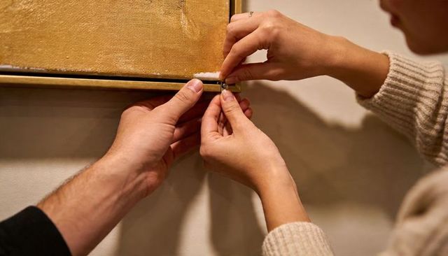 Hands fastening d-ring on gold frame while aligning artwork for wall mounting