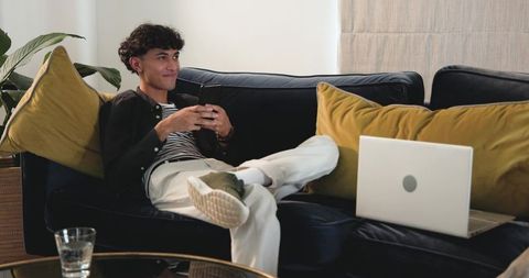 Young man reclining on navy velvet sofa using smartphone with laptop and mustard pillows
