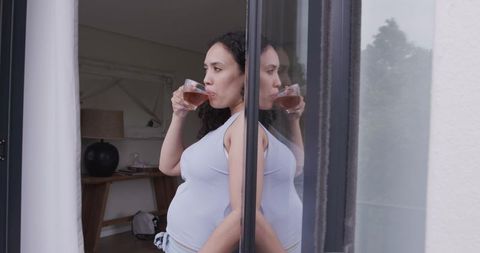 Pregnant african american woman sipping tea on balcony, leaning by sliding glass door reflection