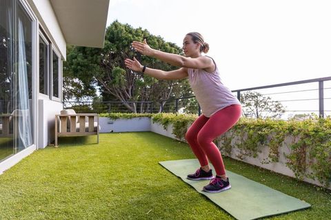 Woman squatting on balcony yoga mat exercising on home terrace with green lawn and sunlight