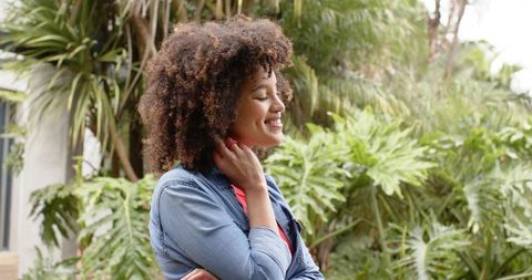 Smiling woman standing with eyes closed touching neck in lush tropical courtyard
