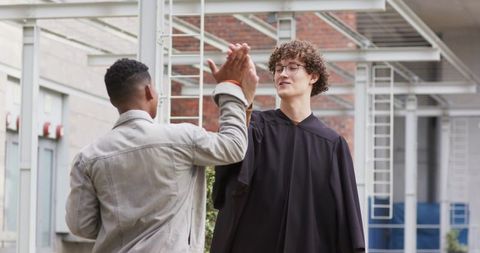 Graduating student high-fiving friend under covered campus walkway wearing academic gown
