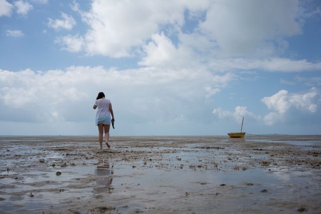 Woman walking barefoot on sandy shore during low tide with a distant boat and clouds in background. Could be used for depicting solitude, reflection, or connection with nature. Ideal for travel blogs, nature magazines, or stress-relief promotions.