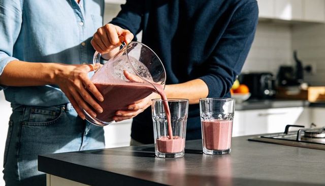 Couple pouring berry smoothie from clear pitcher into tall glasses on modern kitchen island