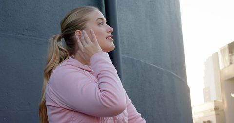 Young woman listening on rooftop in pink jacket looking toward city skyline at golden hour