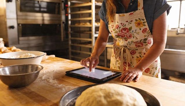 Baker in floral apron using tablet while preparing dough in sunlit artisan bakery