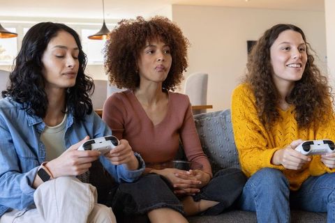 Diverse women playing video game on sofa holding wireless controllers and cheering