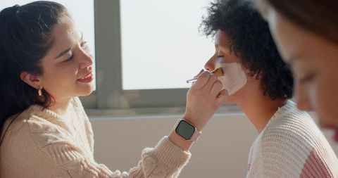 Multiracial women applying facial mask with brush during natural daylight skincare session