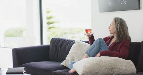 Mature woman relaxing on modern sofa holding tea cup near bright window, minimalist home calm