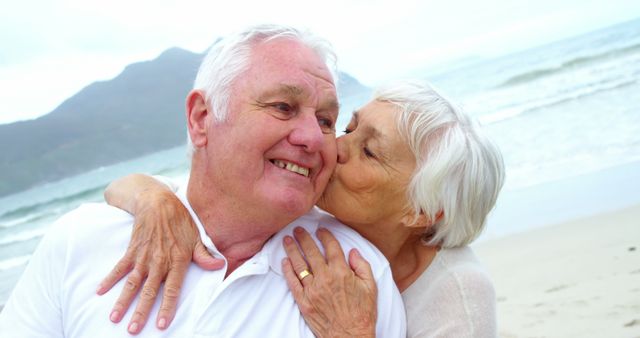 Senior couple showing affection on beach near ocean with mountains in background. Smiling elderly man being kissed by wife, symbolizing love and happiness in retirement. Perfect for campaigns related to retirement living, senior health, romantic vacations, and happy aging.
