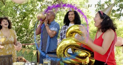 Laughing friends celebrating on sunlit backyard wearing party hats with gold 8 balloon