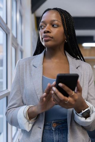 Confident african american businesswoman standing by window holding smartphone