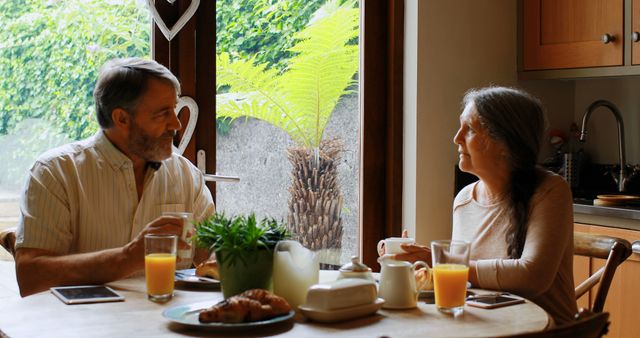 Senior couple sitting at kitchen table enjoying breakfast together, with coffee and orange juice, by window letting in natural light. Useful for articles on aging, living well, retirement lifestyle, and promoting family time, home trends, or kitchen decor.