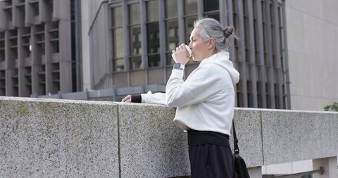 Senior woman sipping coffee while leaning on city concrete railing wearing white hoodie