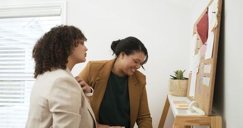 Diverse women adjusting blazers reviewing swatches on corkboard in creative studio