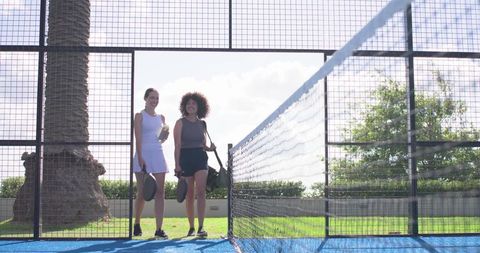 Diverse women walking on blue padel court carrying paddles and bag, smiling after match