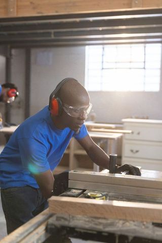 African american carpenter guiding lumber through table saw wearing safety gear