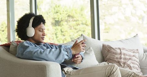 Mid-30s man relaxing on sofa wearing white headphones and using smartphone at home