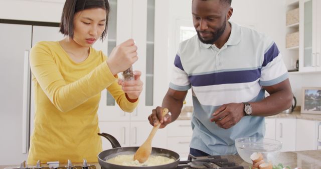 This stock photo depicts a happy, diverse couple enjoying the process of cooking breakfast together in a modern home kitchen. The scene shows both individuals actively participating in meal preparation, embodying themes of teamwork, love, and domestic harmony. The diverse ethnic backgrounds of the couple emphasize inclusivity and cultural acceptance. This image is perfect for websites, blogs, and advertisements that focus on relationship tips, cooking tutorials, morning routines, family-centric brands, kitchen appliances, and lifestyle articles aimed at promoting quality time spent with loved ones.