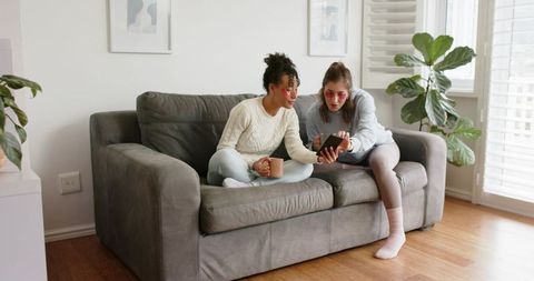 Multiracial friends relaxing on gray couch, sharing smartphone and coffee, cozy home moment