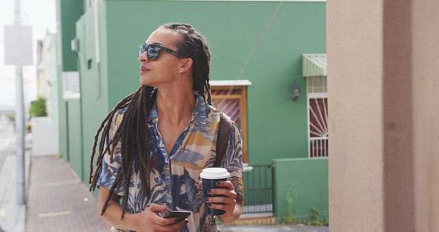 Fashionable biracial man with dreadlocks holding smartphone and takeaway coffee, walking on street. Street style, modern urban lifestyle and communication.